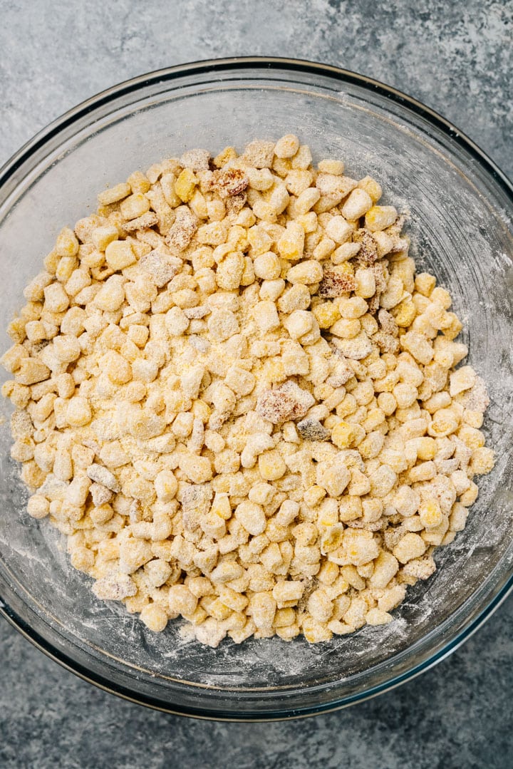 Corn kernels tossed with cornmeal, baking powder, and seasoning in a large glass mixing bowl.