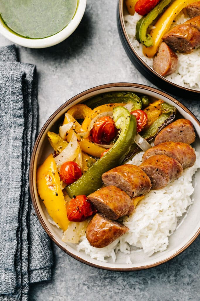 Sheet pan sausage and peppers over white rice in a bowl with a black linen napkin and bowl of basil sauce on the side.