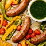Cooked sausages, peppers, and tomatoes on a sheet pan with a small bowl of basil sauce on the side.