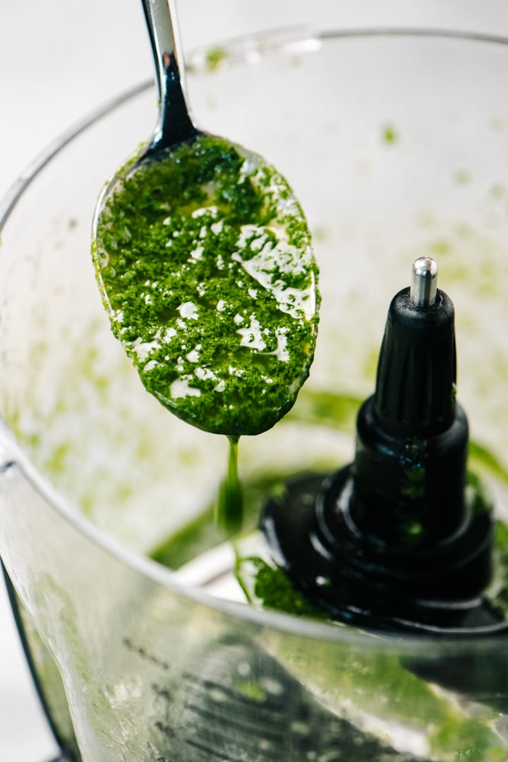 Basil sauce dripping from a spoon hovering over a food processor.