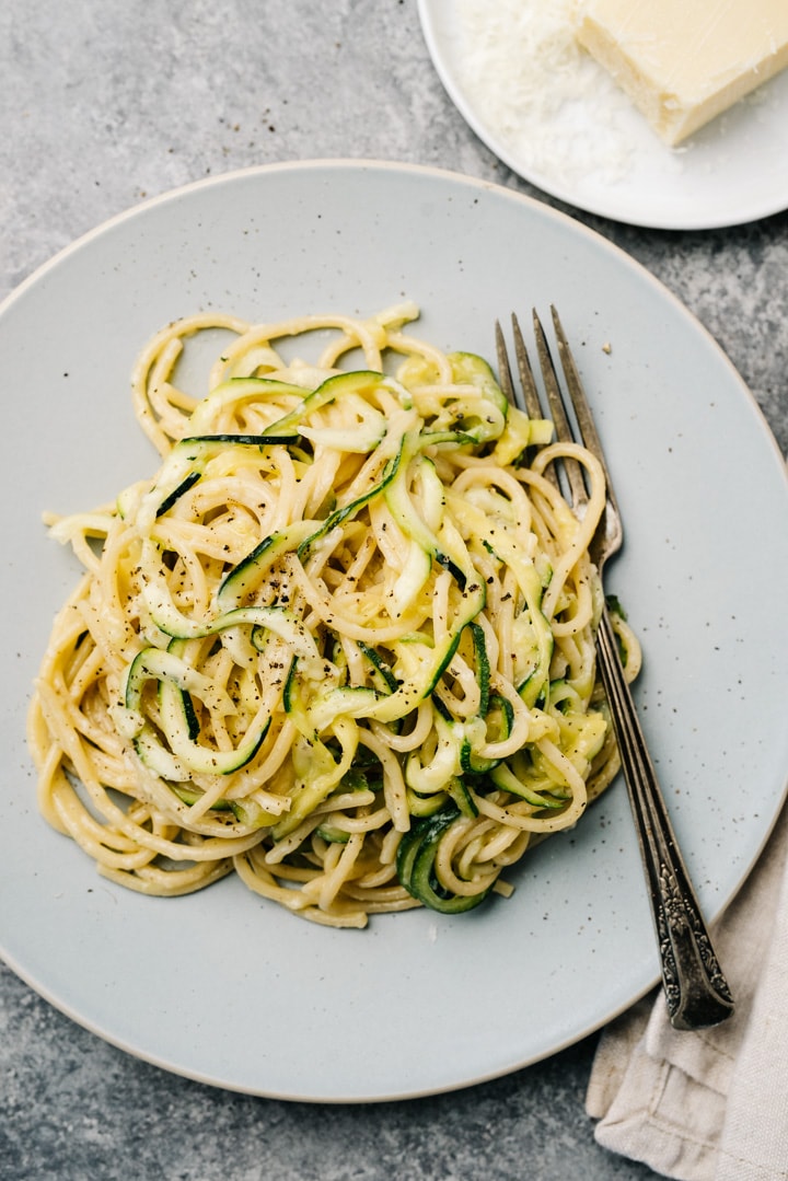 Cacio e pepe pasta with zucchini noodles on a blue plate with a fork, and a small dish of freshly grated parmesan cheese to the side.