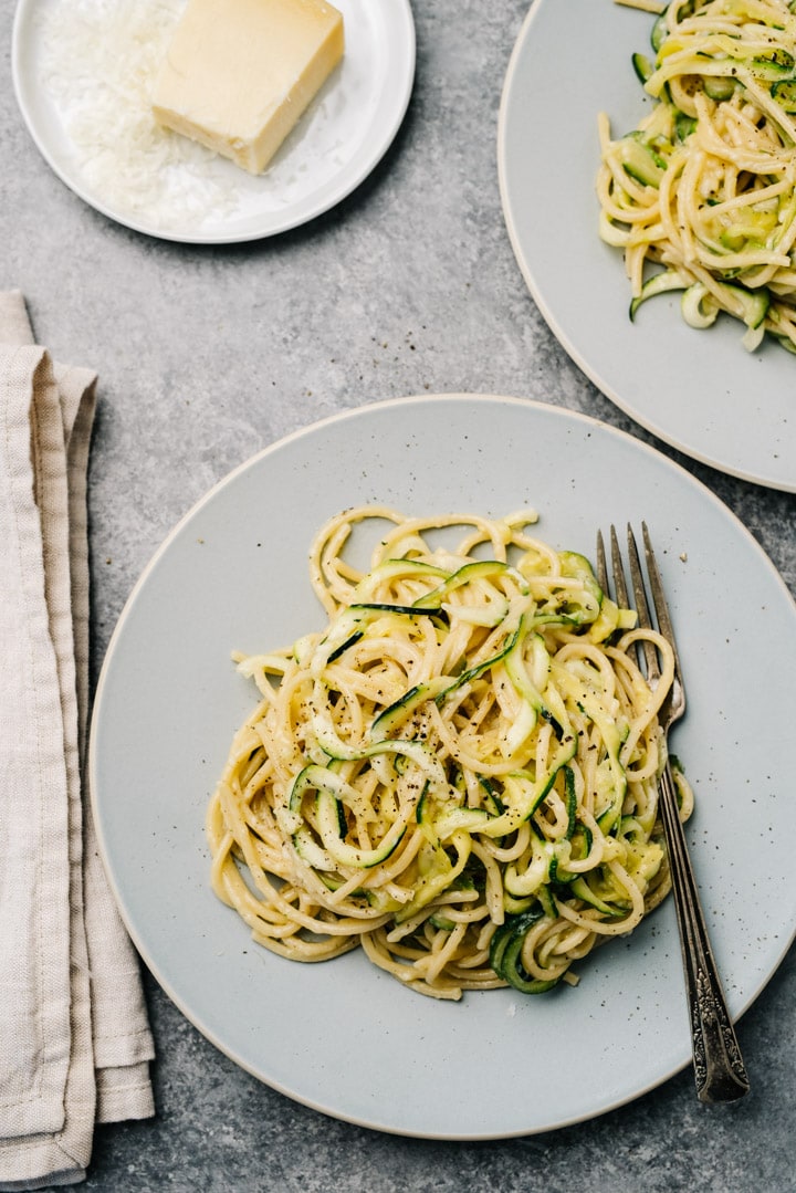 Two plates of zucchini pasta on a cement background with a linen napkin and fresh parmesan cheese to the side.