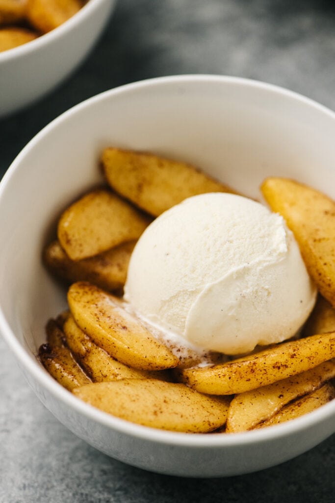 Side view, air fried apples in a white bowl topped with a scoop of vanilla ice cream on a concrete background.