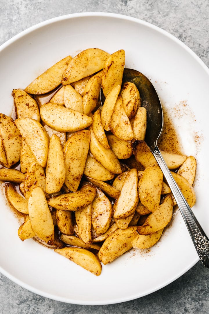 Air fryer apples tossed with brown sugar and maple syrup in a white bowl with a silver serving spoon.