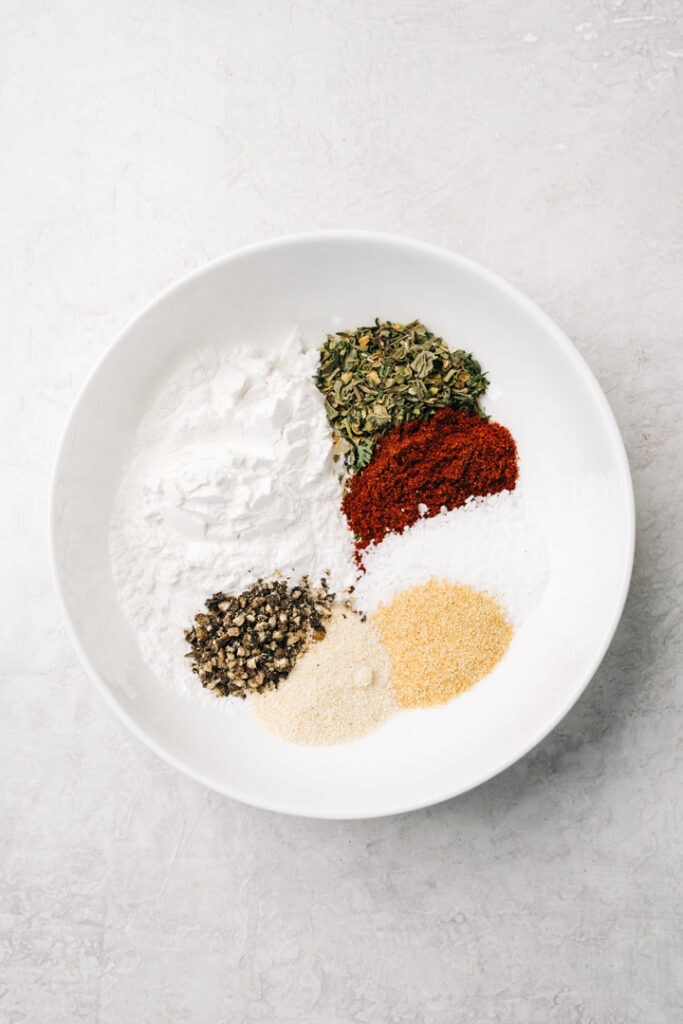 The ingredients for chicken drumsticks dry rub in a small white bowl - baking powder, italian seasoning, paprika, salt, garlic powder, onion powder, and pepper.