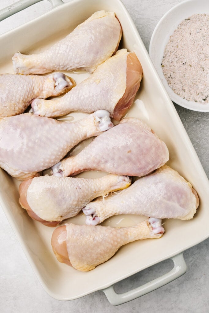 Chicken drumsticks arranged in a casserole dish with a bowl of dry rub to the side.