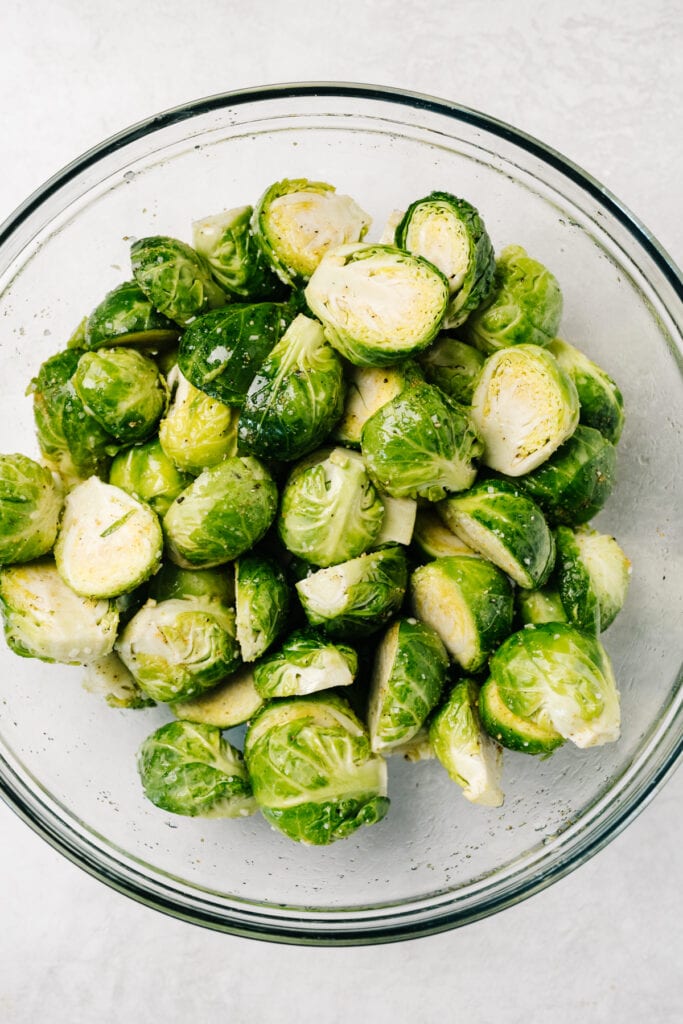 Halved brussels sprouts in a glass mixing bowl, drizzled with olive oil and seasonings.