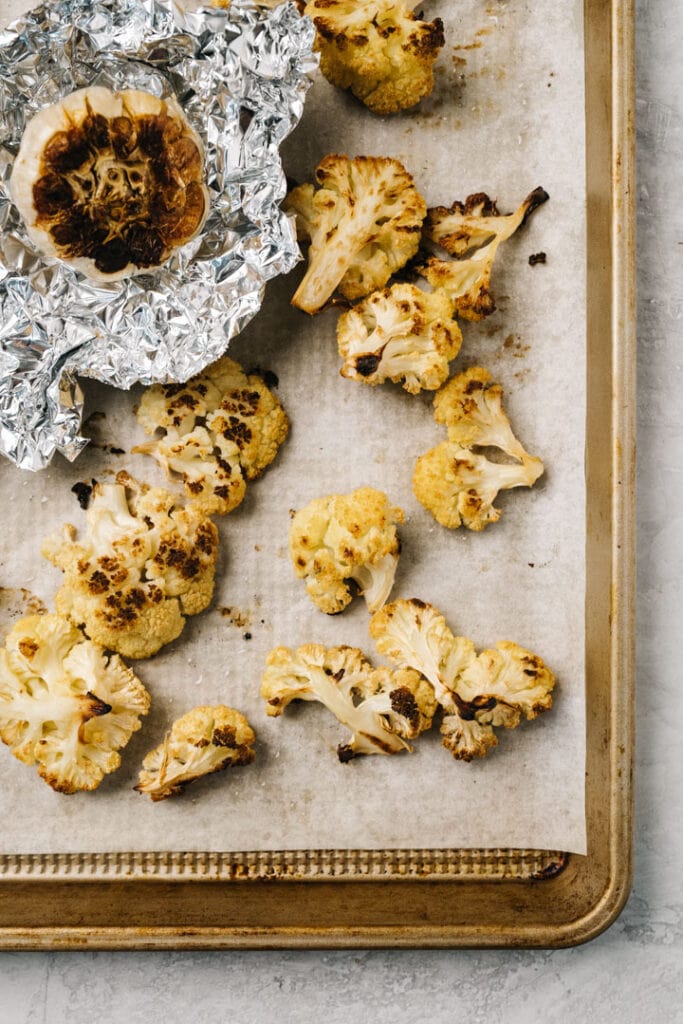 Roasted cauliflower florets and roasted garlic on a parchment lined baking sheet.