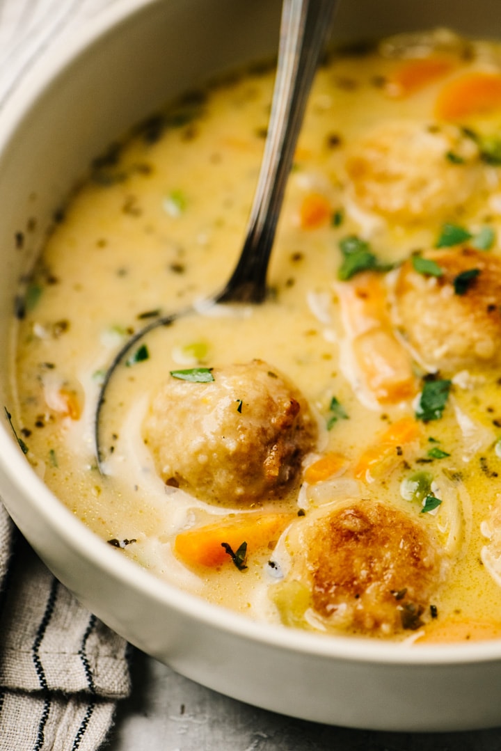 Side view, a silver soup spoon tucked into a bowl of creamy chicken meatball soup.