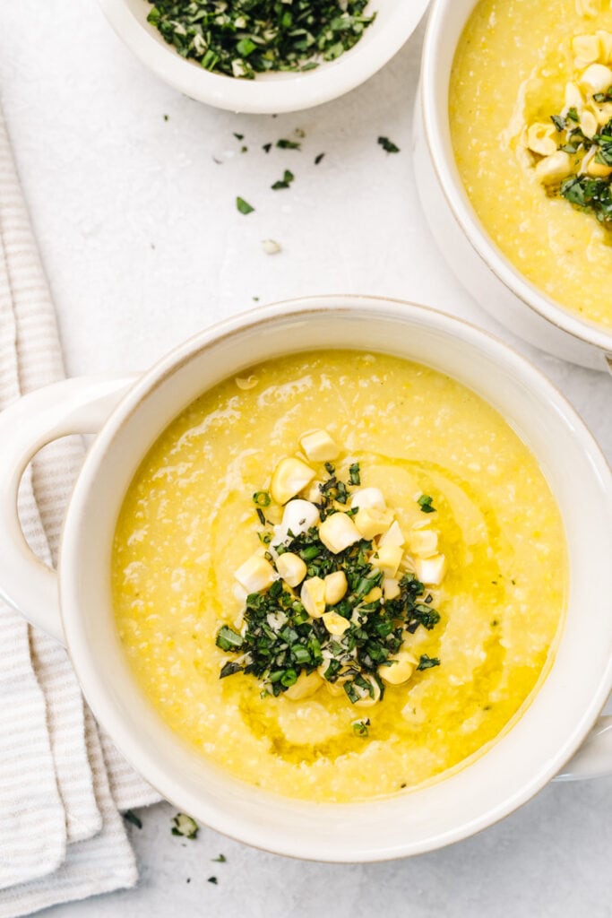 Two bowls of healthy corn soup on a concrete background with a striped cream napkin and small bowl of gremolata.