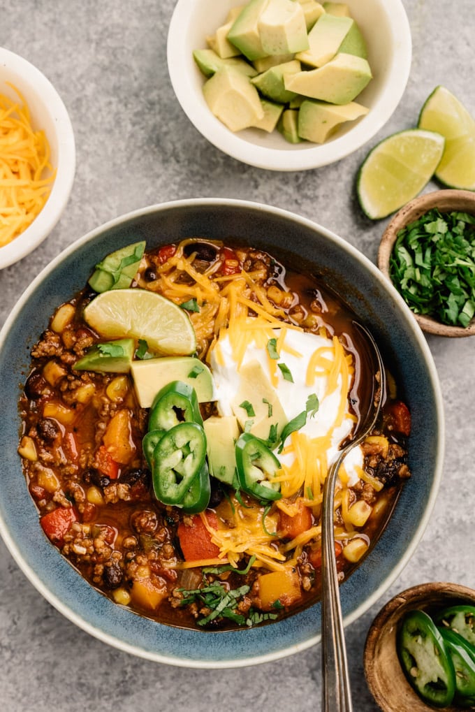 Ground beef taco soup in a blue bowl surrounded by several small bowls of soup garnishes, like avocado, cilantro, cheese, and lime wedges.