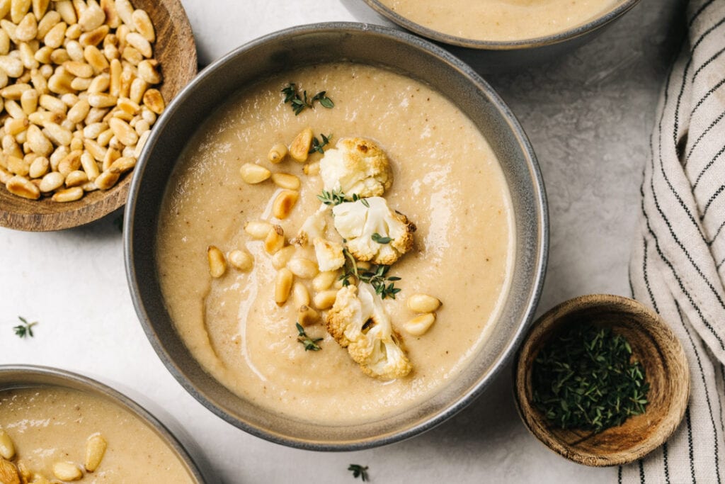 A bowl of creamy homemade cauliflower soup garnished with pine nuts and thyme on a concrete background with a striped linen napkin.