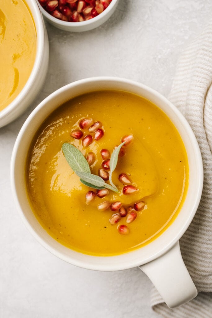A bowl of butternut squash soup in a cream crock, garnished with pomegranate seeds and fresh sage.