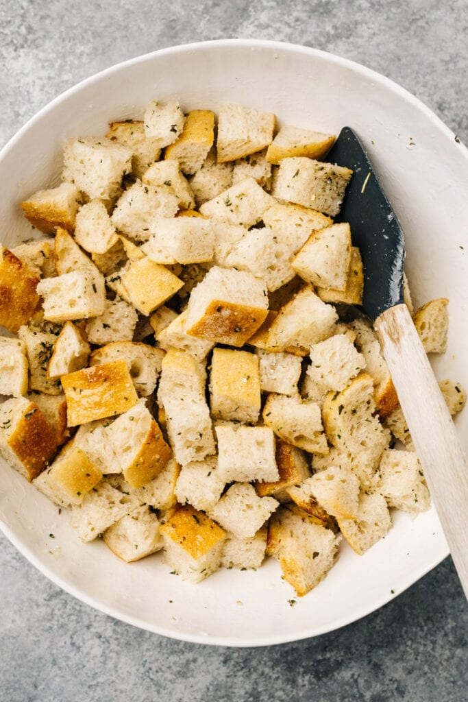 Italian bread cubes tossed with olive oil, salt, pepper, and dried herbs in a large white mixing bowl.