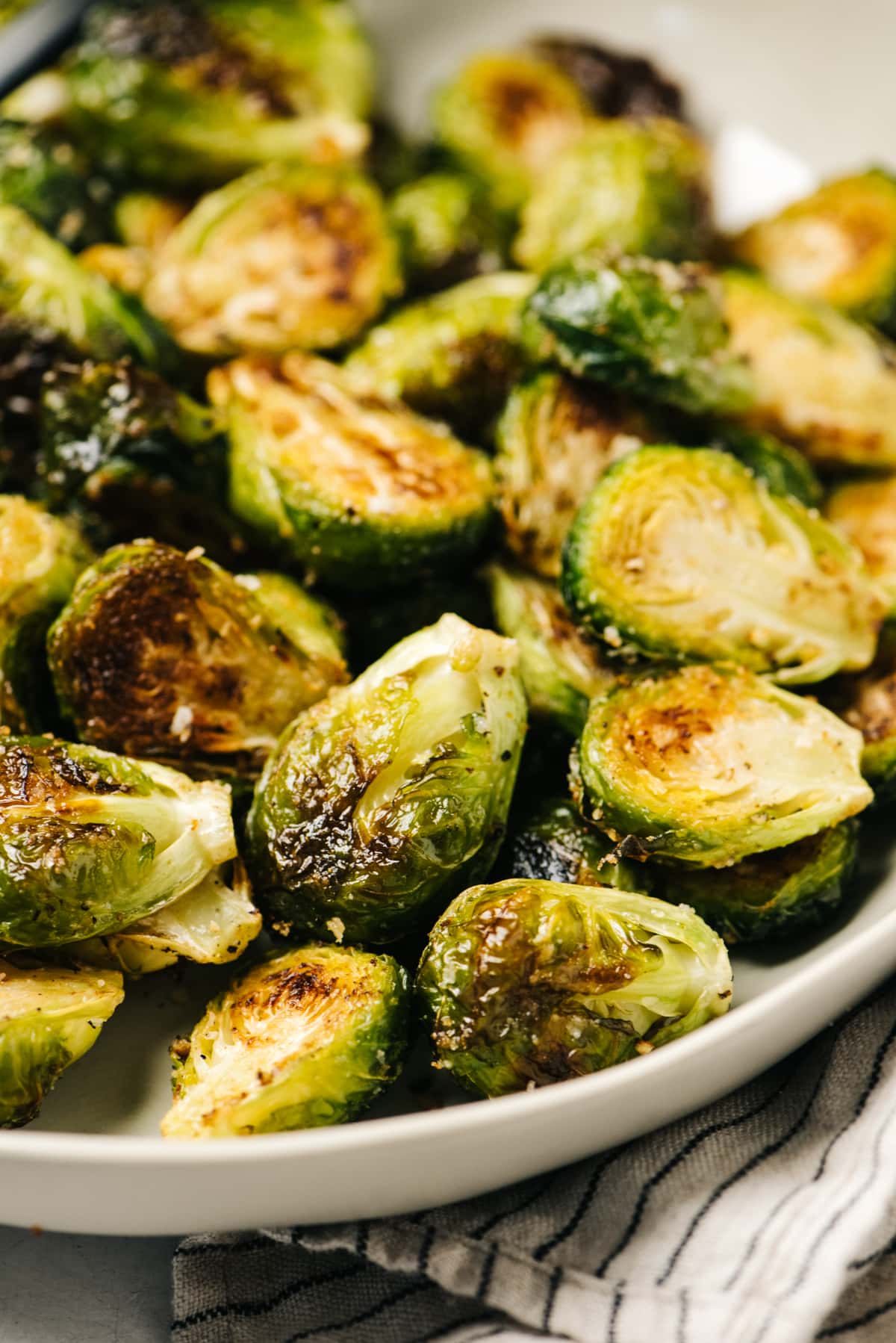 Side view, roasted Brussels sprouts in a low tan serving bowl with a striped napkin tucked under the bowl.