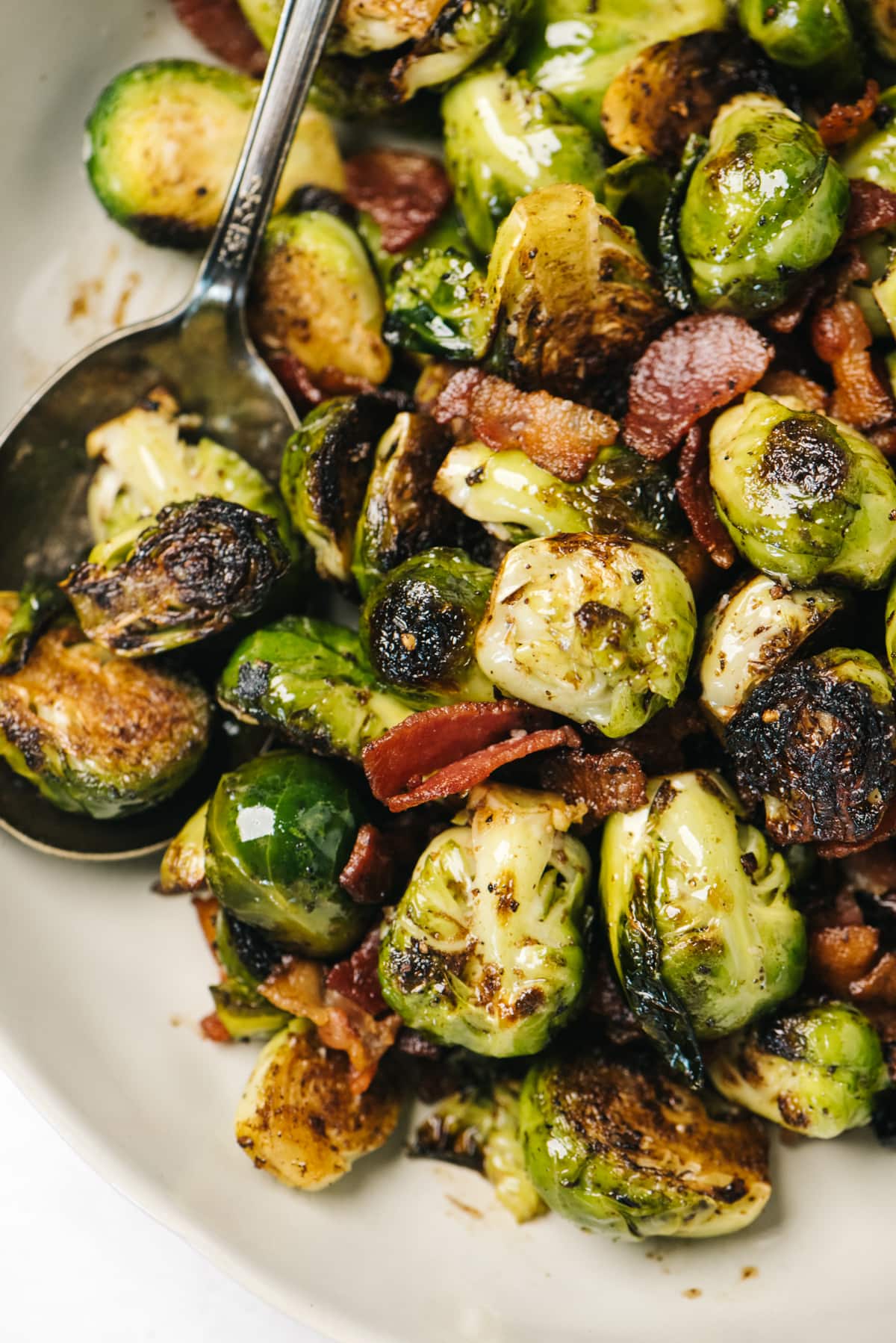 A spoon tucked into crispy sautéed Brussels sprouts with bacon in a low tan serving dish.