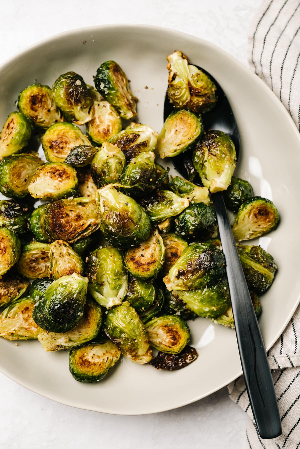 Roasted Brussels sprouts in a low tan serving bowl with a black serving spoon; a striped linen napkin is to the side, partially tucked under the bowl.