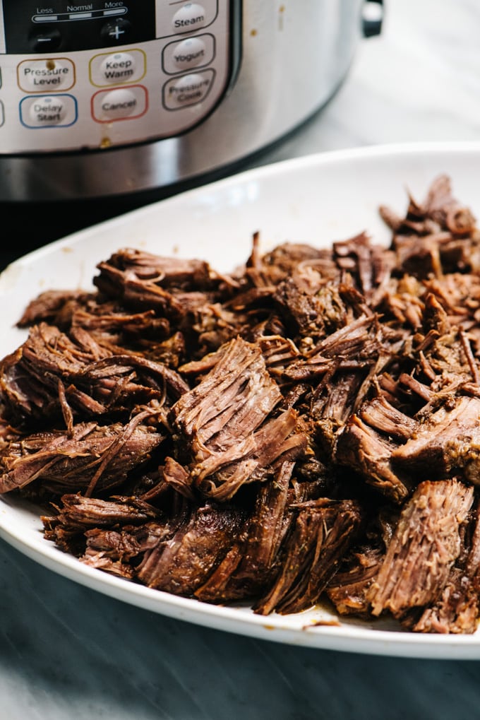 Side view, shredded chuck roast on a white platter with an instant pot in the background.