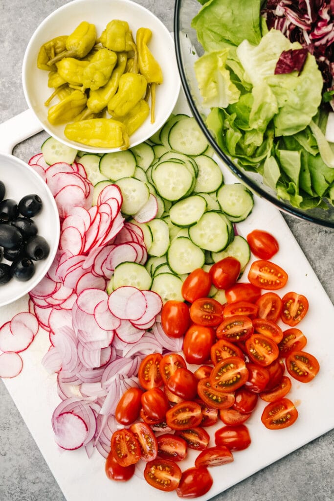 Chopped vegetables arranged on a cutting board - tomatoes, radishes, red onions, cucumber, olives, and pepperoncini.