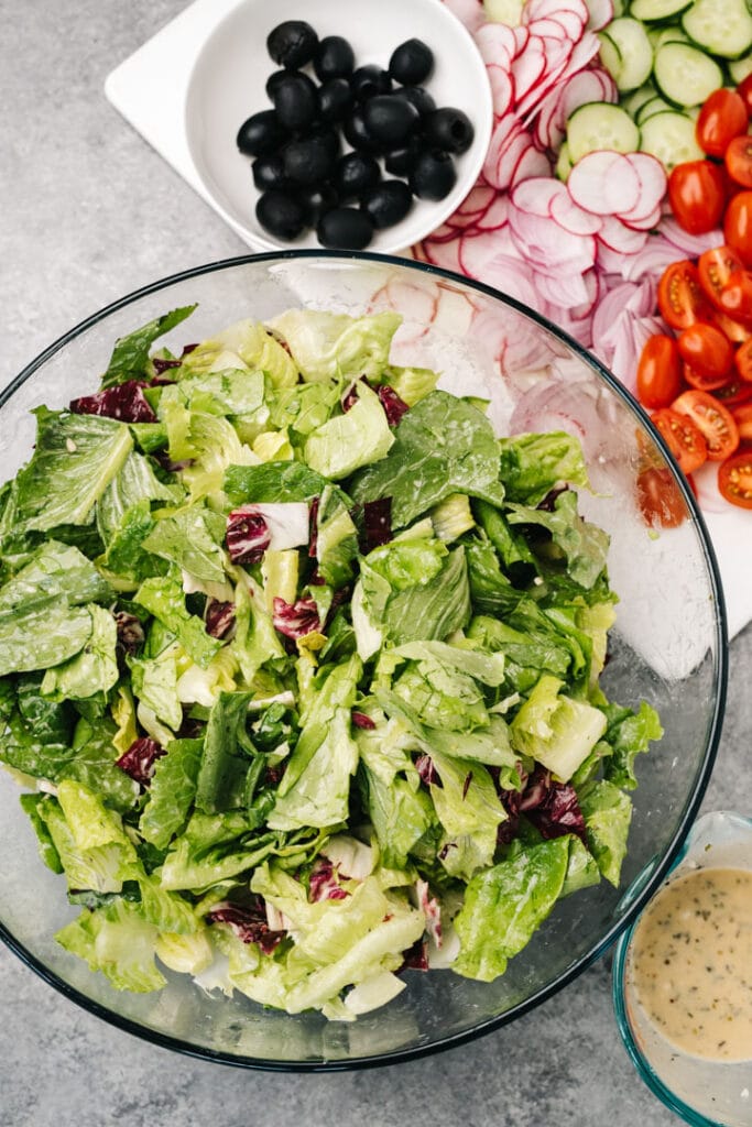 Chopped romaine lettuce and radicchio tossed with italian dressing in a large mixing bowl with sliced vegetables on a cutting board to the side.