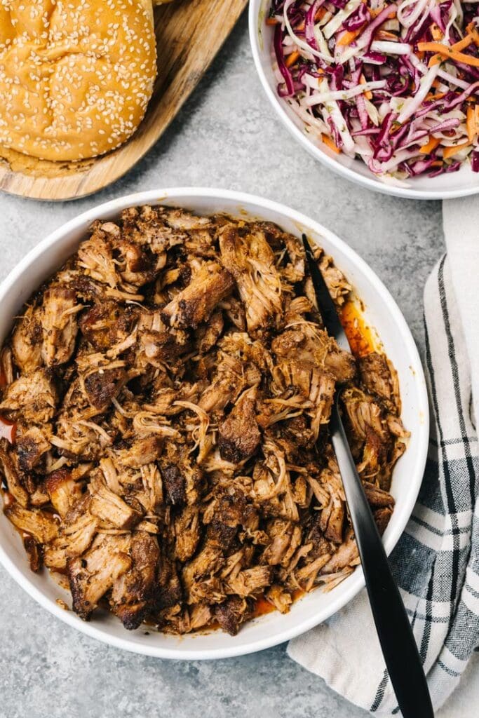 Dutch oven pulled pork in a white serving bowl  with a black serving fork surrounded by brioche buns, coleslaw, and a linen kitchen towel.