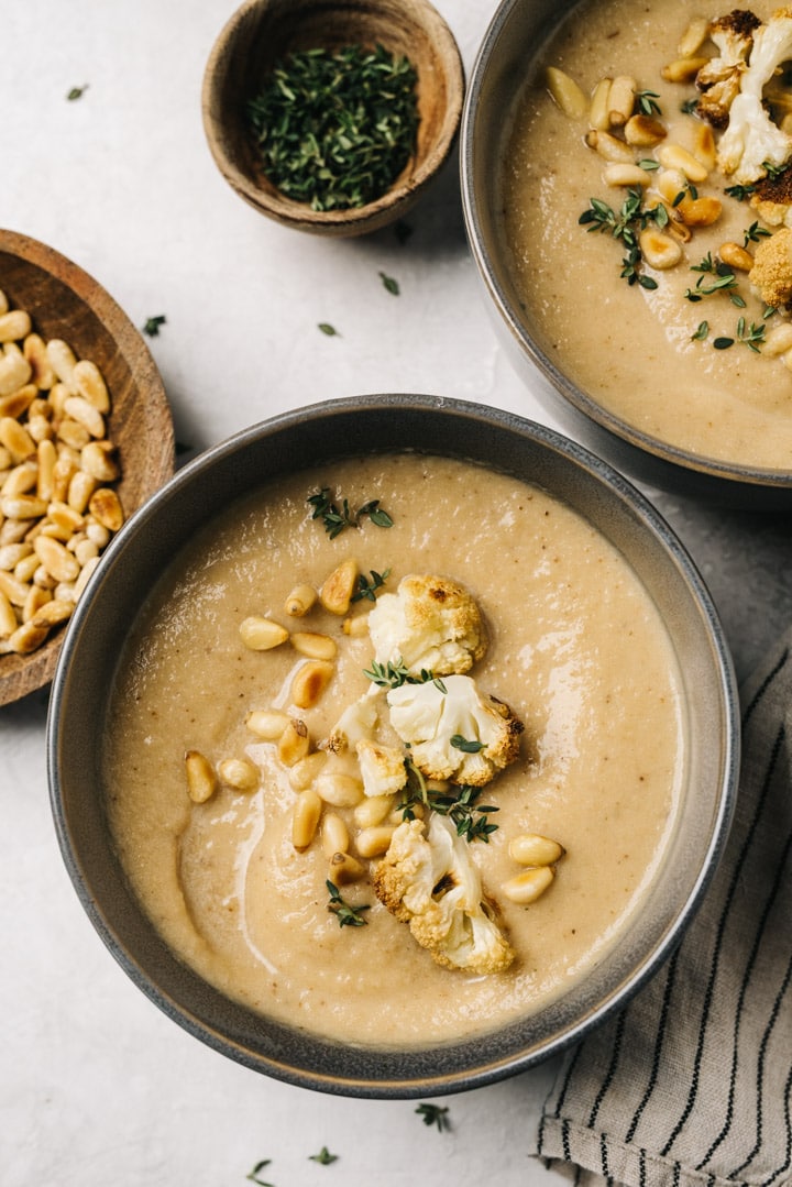 Two bowls of roasted cauliflower soup on a cement background with small bowls of pine nuts and thyme and a striped linen napkin to the side.