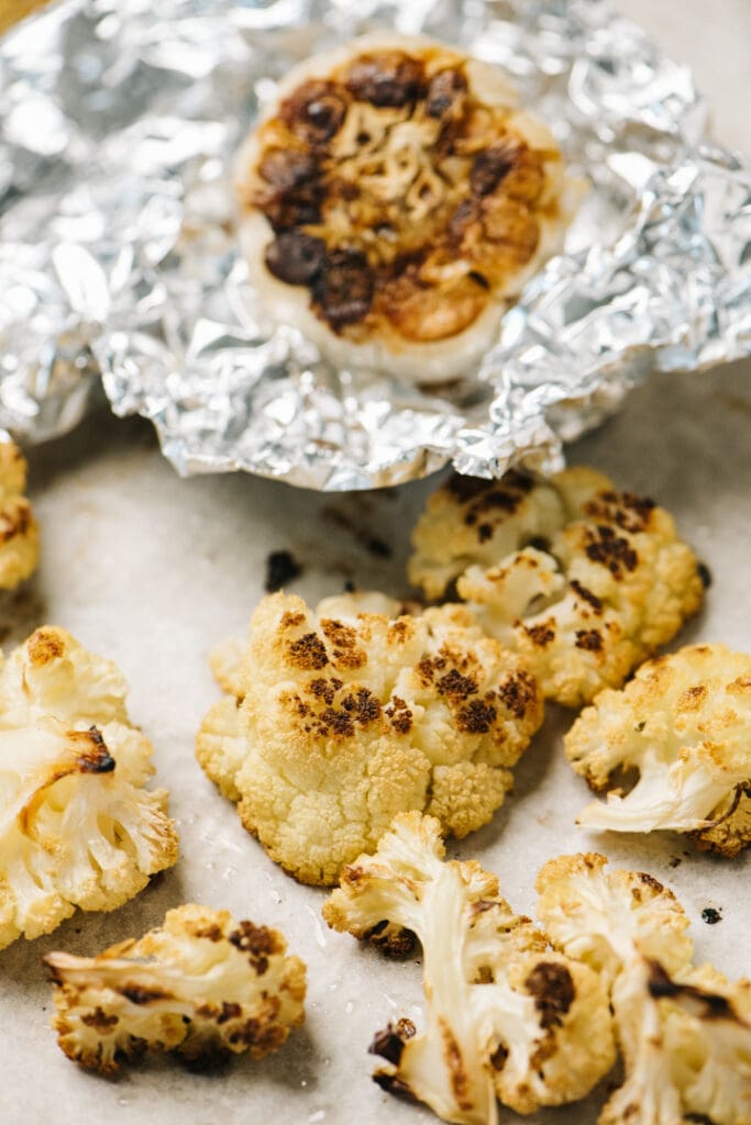 Side view, roasted cauliflower florets on a baking sheet with a head of roasted garlic in the background.
