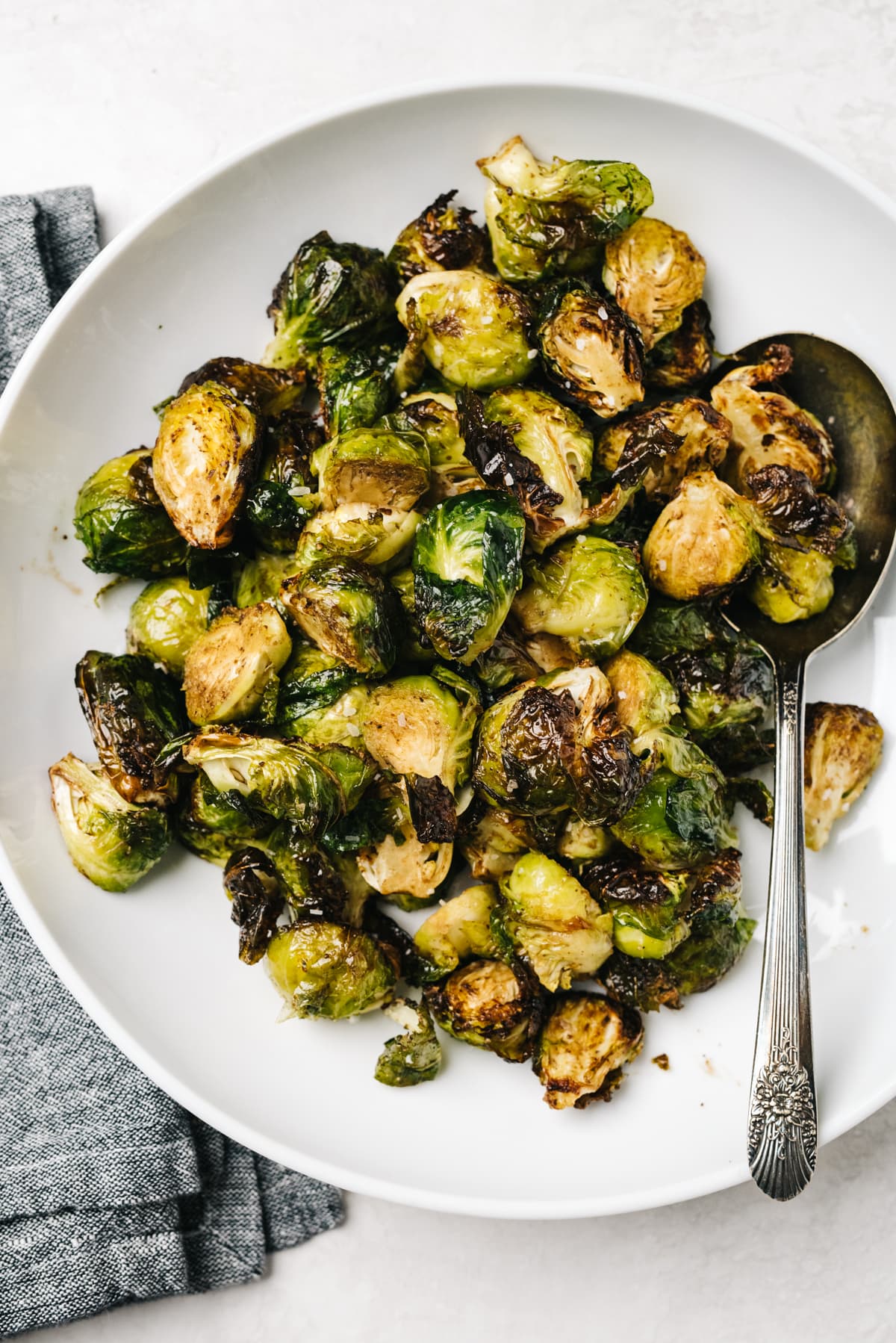 Roasted air fried brussels sprouts in a white serving bowl with a silver serving spoon and dark grey linen napkin on the side.