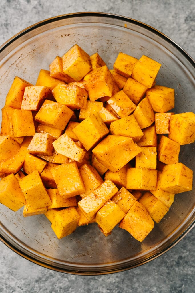 Diced butternut squash tossed with olive oil, salt, cinnamon, and nutmeg in a glass mixing bowl.