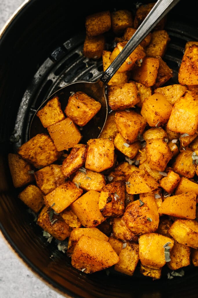 Air fryer butternut squash with maple, cinnamon, and sage in an air fryer basket with a silver serving spoon.