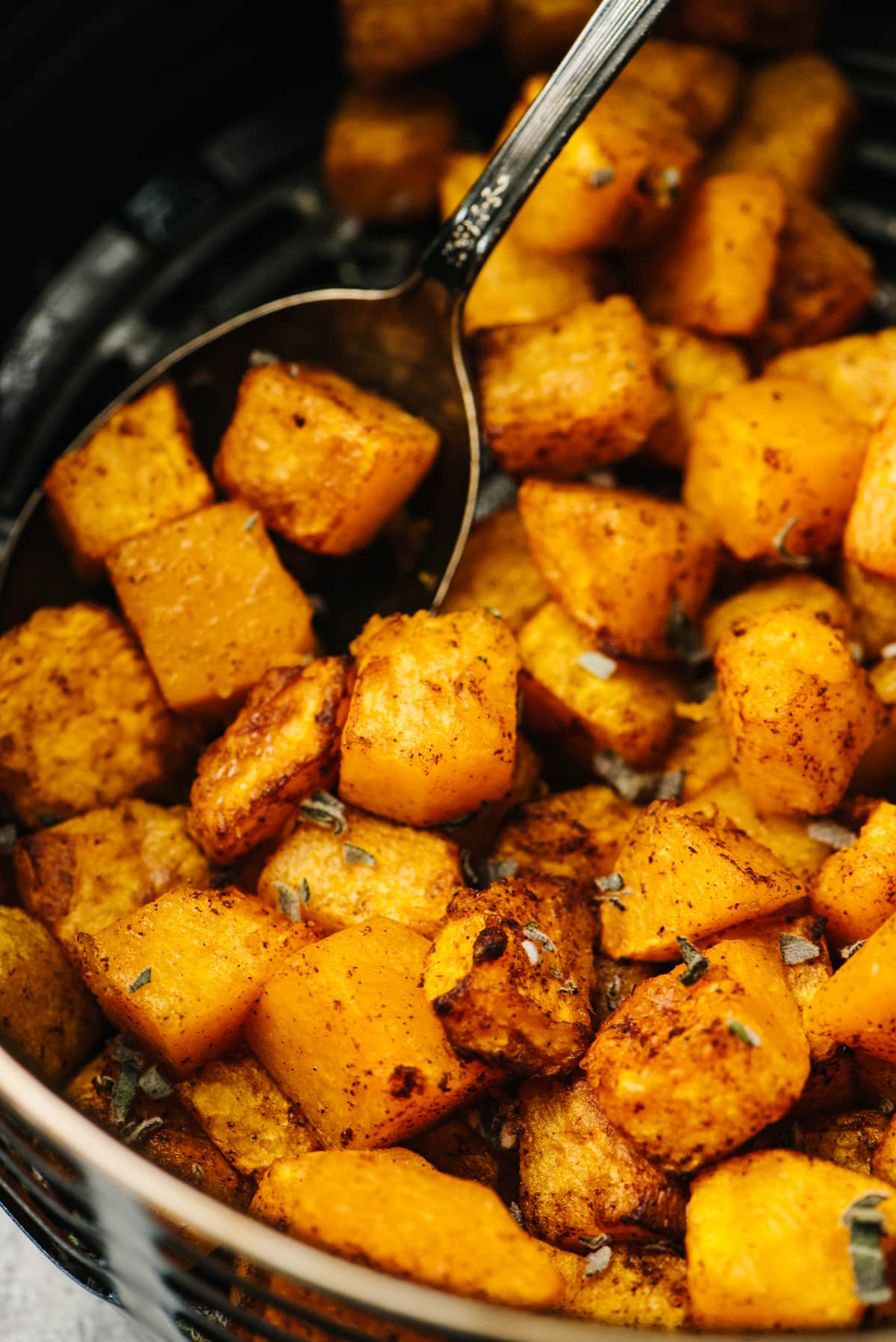 Roasted diced butternut squash in the basket of an air fryer with a vintage serving spoon, garnished with fresh sage.