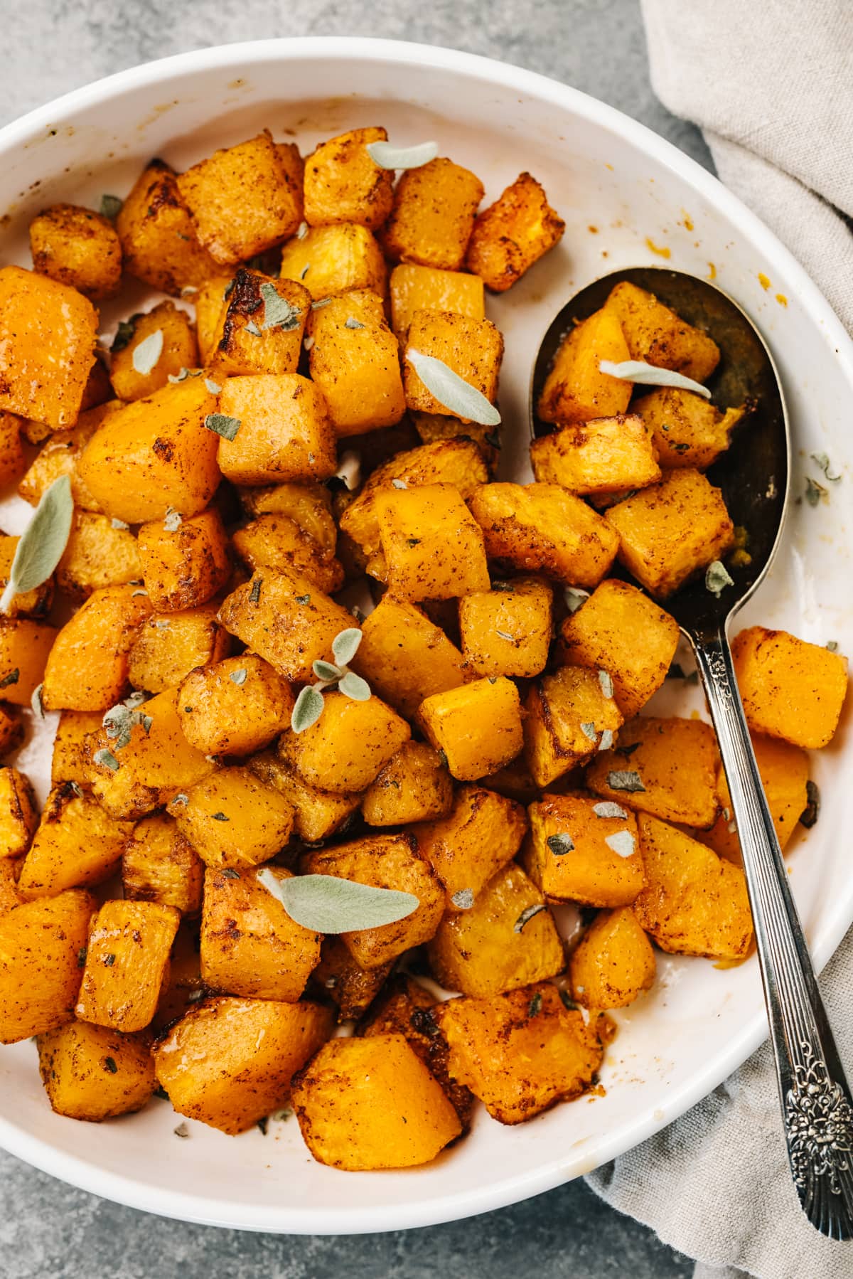 Air fryer butternut squash cubes in a white serving bowl with a vintage serving spoon and white linen napkin to the side.