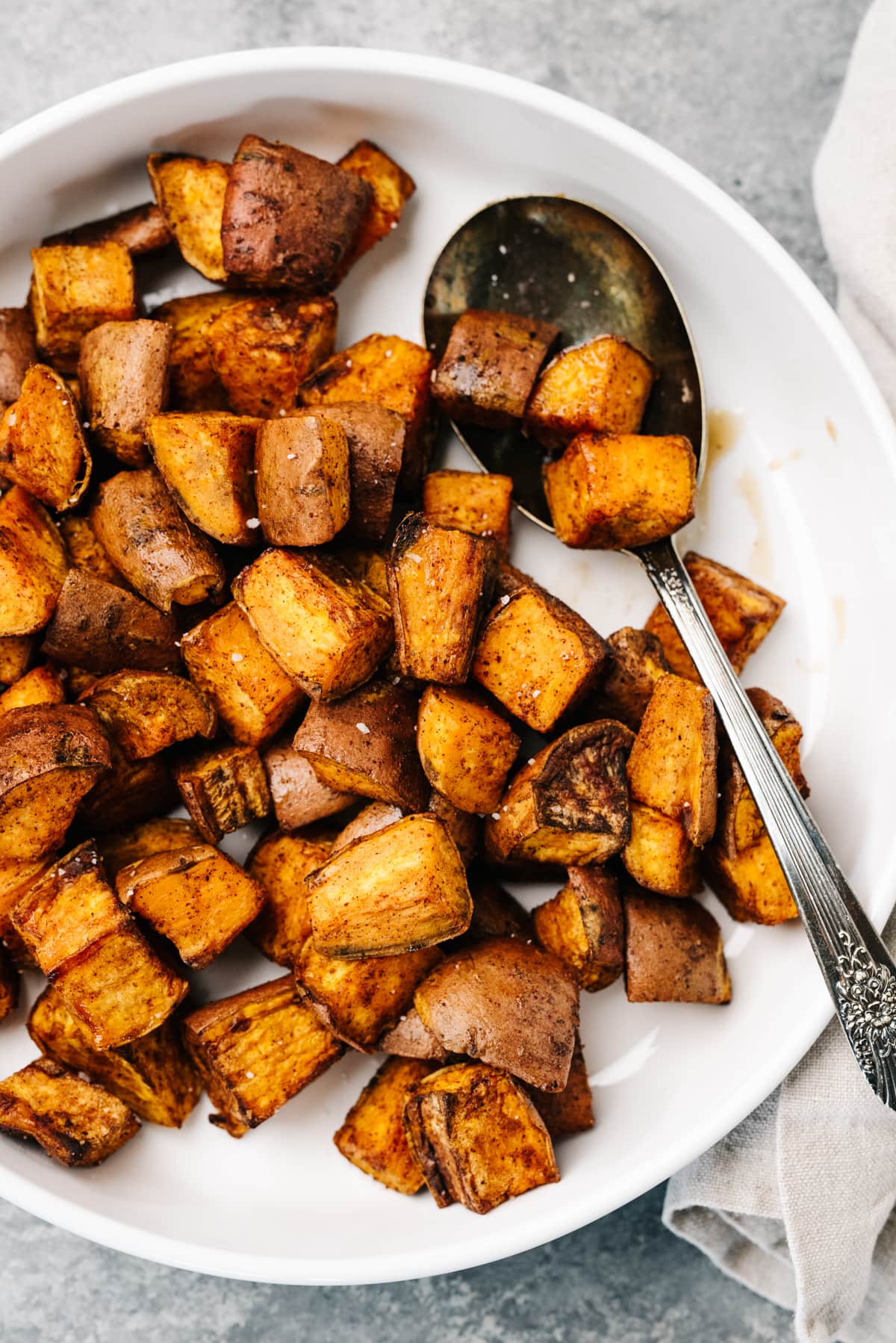 Air fryer sweet potatoes in a white serving bowl with a serving spoon and tan linen napkin to the side.