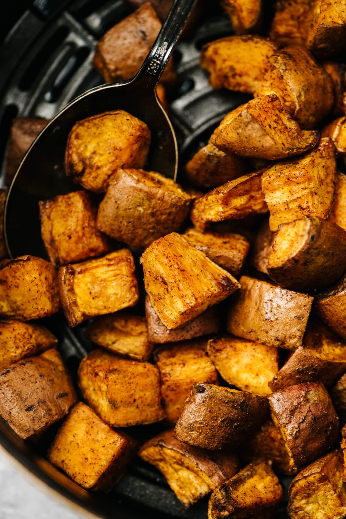 A silver serving spoon tucked into roasted sweet potatoes in the basket of an air fryer.