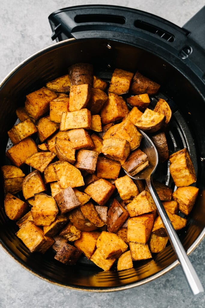 A spoon tucked into sweet potato cubes in an air fryer, showing to stir halfway through cooking.