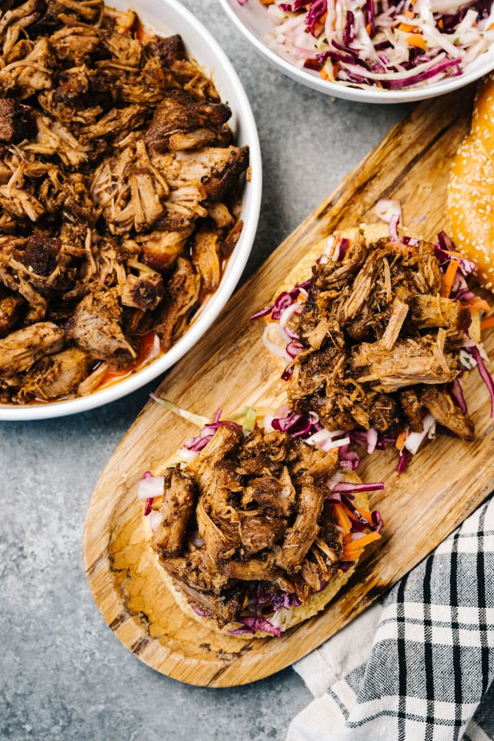 Two pulled pork sandwiches on a wood cutting board with a serving dish of coleslaw and dish of pulled pork to the side.