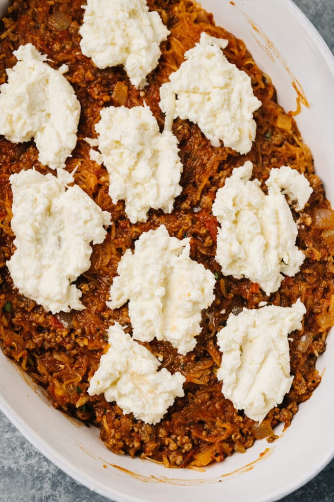 A layer of spaghetti squash lasagna in a casserole dish - a layer of squash, meat, and tomato sauce, topped with dollops of a ricotta cheese mxiture.