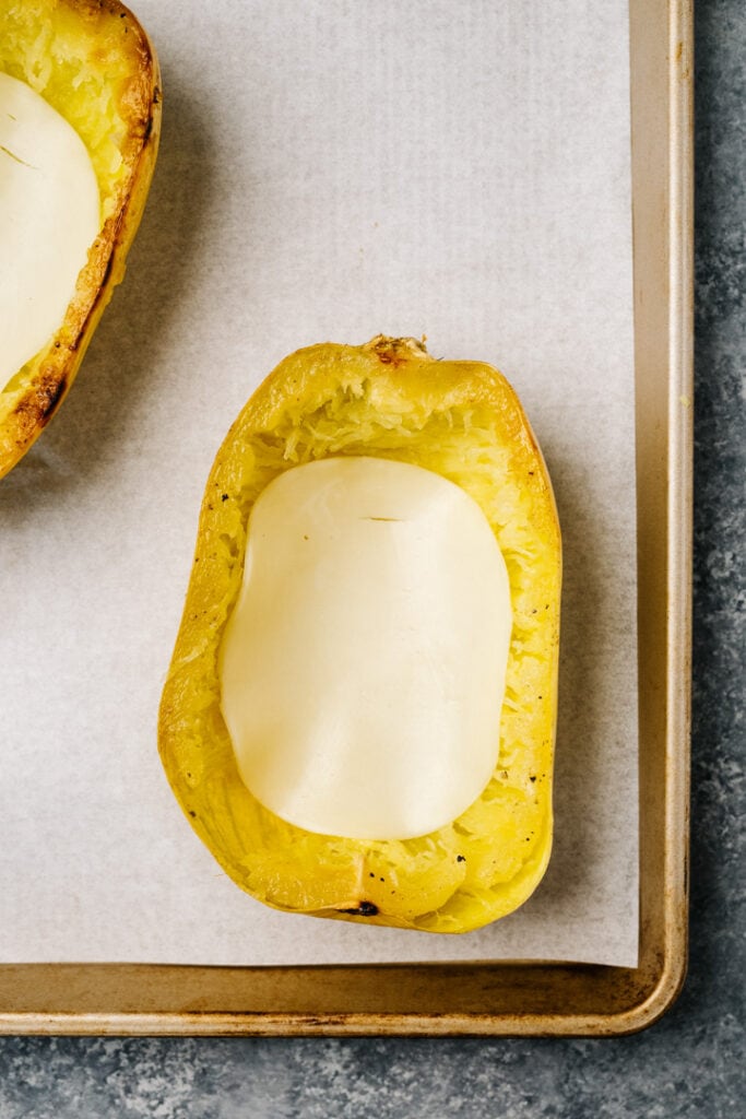 Roasted spaghetti squash halves lined with provolone cheese on a parchment lined baking sheet.