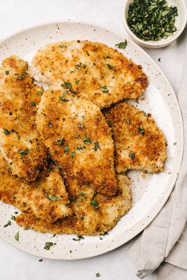 Baked parmesan crusted chicken breasts on a speckled tan serving platter, garnished with parsley; a tan linen napkin and small bowl of parsley to the side.