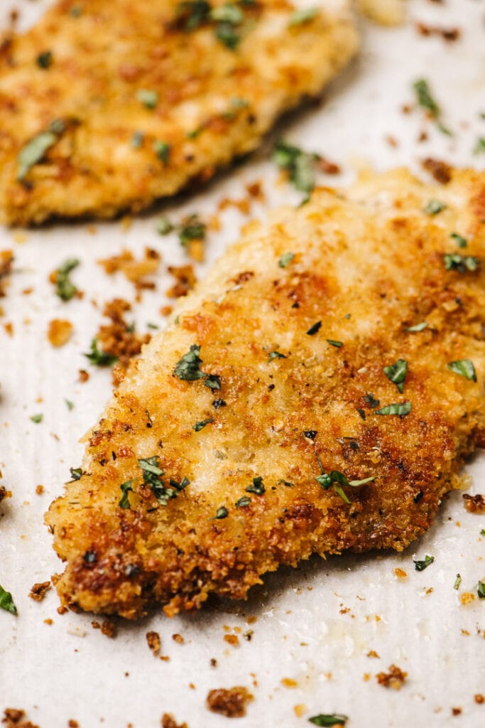 Side view, two golden brown parmesan crusted chicken breasts on a parchment lined baking sheet, garnished with fresh chopped parsley.