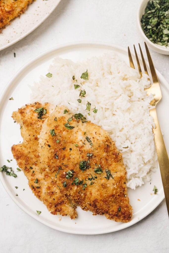 Two baked parmesan crusted chicken breasts over steamed white rice on a white plate with a gold fork.