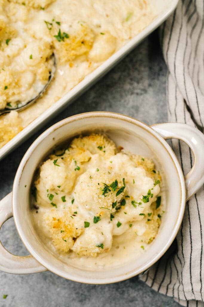 Cauliflower au gratin in a small crock bowl next to a casserole dish with a linen napkin to the side.