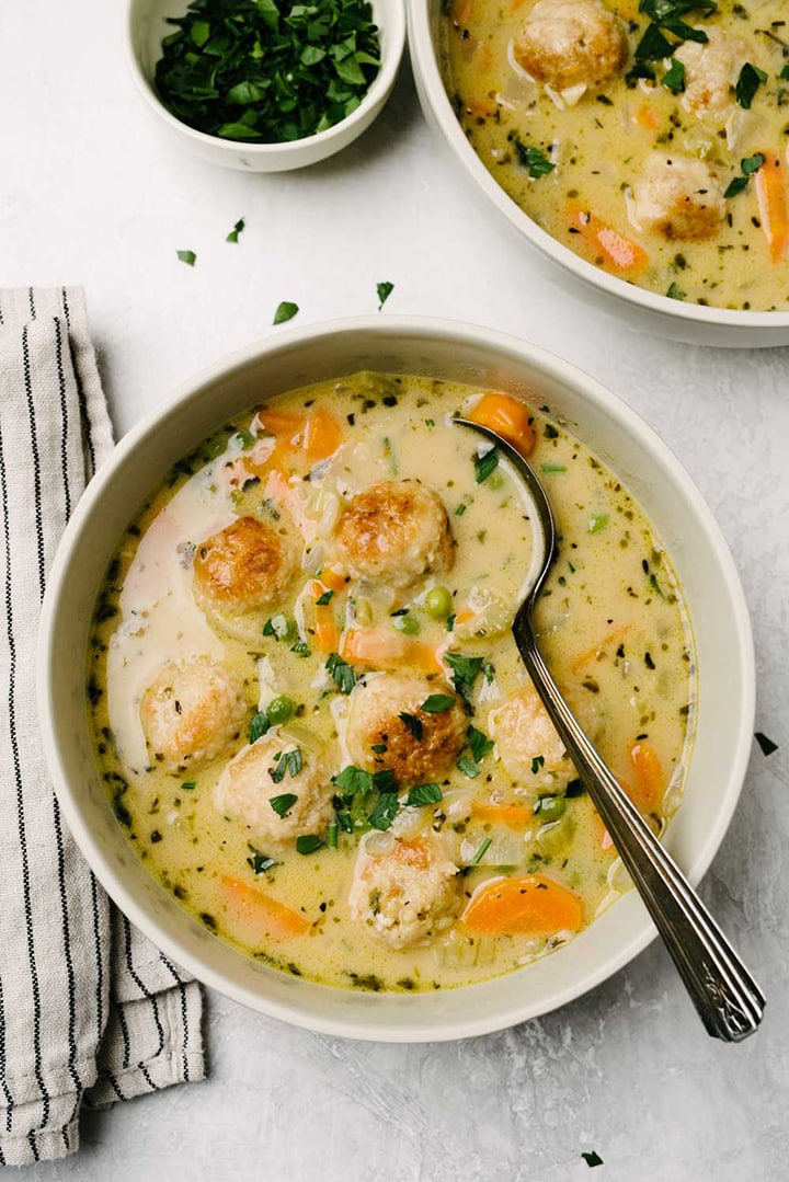 A bowl of creamy chicken soup made with mini chicken meatballs on a concrete background with a striped linen napkin and bowl of minced parsley garnish.