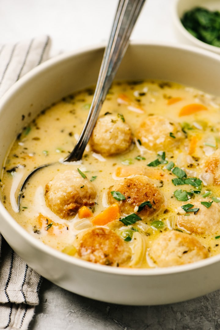 Side view, a bowl of creamy chicken meatball soup in a low tan bowl with a silver soup spoon.