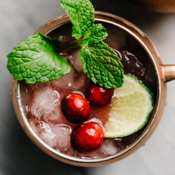 From overhead, a cranberry Moscow mule cocktail in a copper mug on a marble table, garnished with a lime wedge, mint sprig, and fresh cranberries.