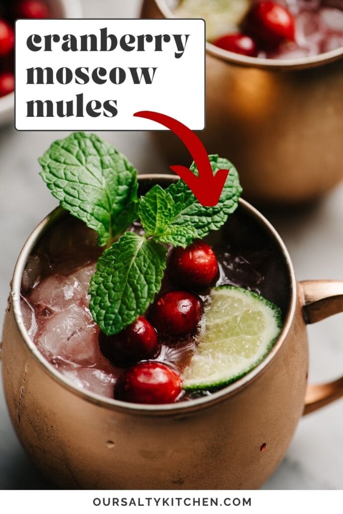 Side view, a Moscow mule with homemade cranberry syrup in a copper mug, garnished with a lime wedge, mint sprig, and fresh cranberries; a second cocktail and a small bowl of cranberries are in the background; text overlay reads "cranberry moscow mules".