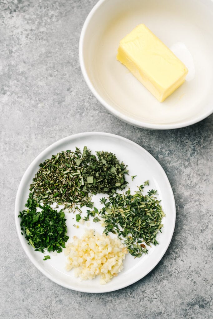 Room temperature butter, chopped fresh herbs, and minced garlic arranged on a concrete background.