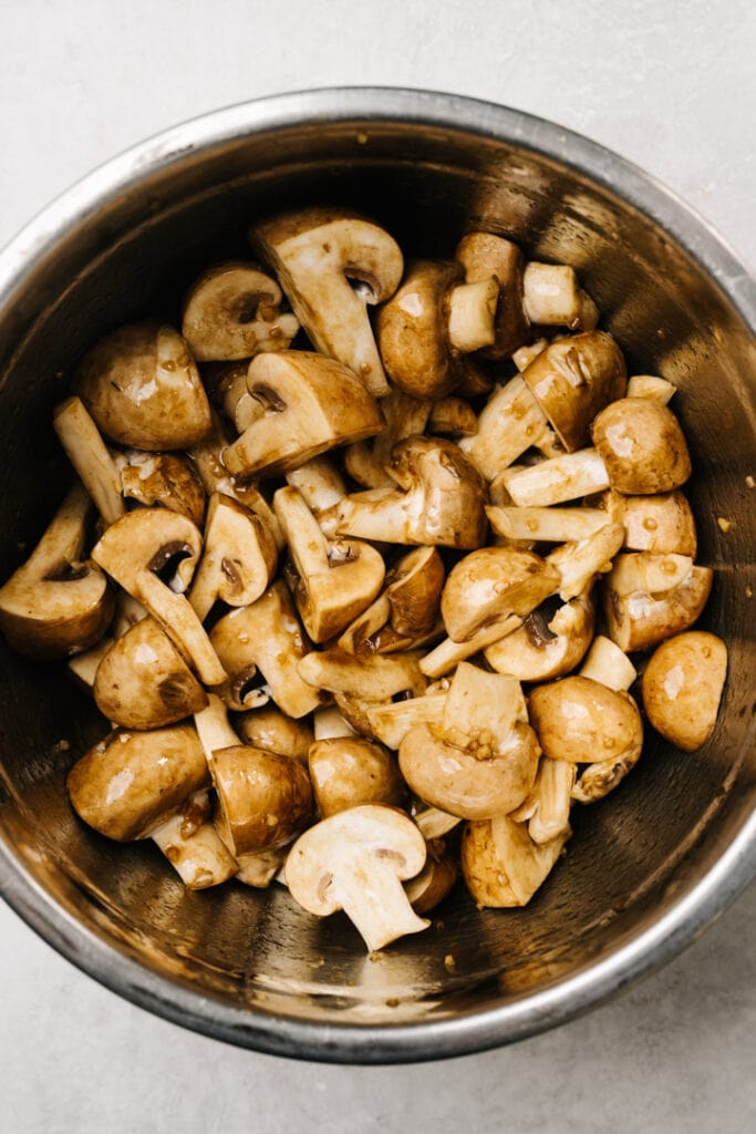 Quartered crimini mushrooms tossed with a mixture of butter, olive oil, balsamic vinegar, soy sauce, and minced garlic in a metal mixing bowl.