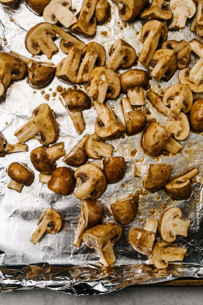 Quartered mushrooms tossed with seasoning on a foil lined baking sheet.