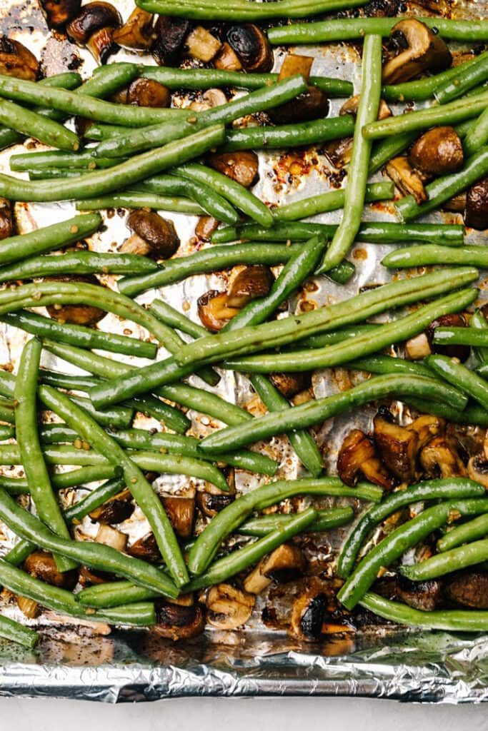 Marinated green beans added to foil lined baking sheet with partially roasted mushrooms.